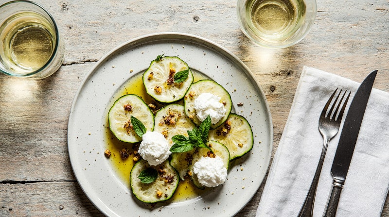 Rezeptidee Wein zum Essen Zucchinisalat mit Ricotta, Pistazien und Zitronenabrieb auf flachem Teller dazu ein Glas Weißwein