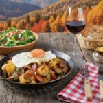 Wein zum Essen mit Südtiroler Bauerngröstel aus Rindfleisch und Kartoffeln in der Pfanne serviert, dazu ein Glas Vernatsch auf rustikalem Holztisch mit Bergblick