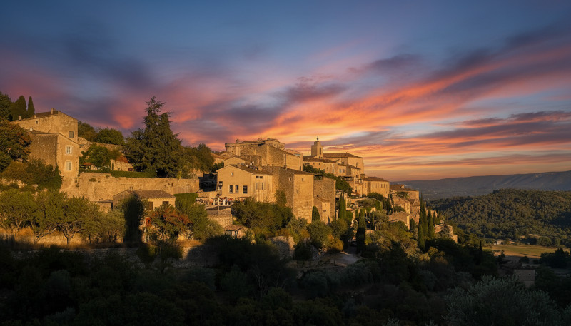 Wein zum Essen – Blick auf das provenzalische Dorf Gordes bei Sonnenuntergang, Natursteinhäuser auf einem Hügel in der Provence, umgeben von Olivenbäumen, warmes Abendlicht und typische Landschaft Südfrankreichs.