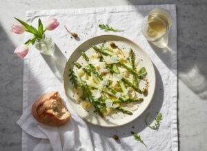 Spargel-Carpaccio roh mit Parmesanspänen und Zitronenöl auf cremefarbener Platte, Pinot Grigio Weißweinglas, norditalienisches Marmor-Setting