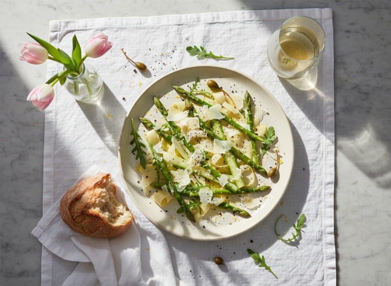 Spargel-Carpaccio roh mit Parmesanspänen und Zitronenöl auf cremefarbener Platte, Pinot Grigio Weißweinglas, norditalienisches Marmor-Setting