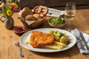 Weißer Spargel mit Kalbsschnitzel und Petersilienkartoffeln auf ovalem Porzellanteller, bayerisches Sonntagslicht, Weißweinglas leicht beschlagen