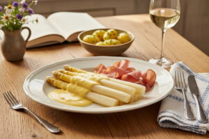 Weißer Spargel mit Sauce Hollandaise und Schwarzwälder Schinken auf ovalem Porzellanteller, bayerisches Sonntagslicht, mit Weißweinglas