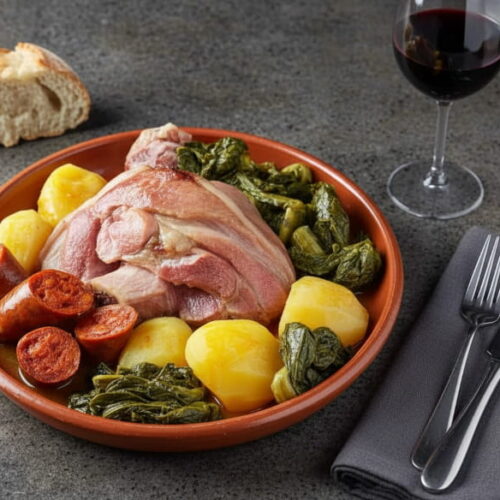 Lacón con grelos auf Tonplatte, Schweinefleisch rosa und saftig, Grelos dunkelgrün, Chorizo rot, Kartoffeln gelbweiß, Rotweinglas mit Mencía dunkelrot, dunkelgrauer Granit, atlantisches Winterlicht, schräge Vogelperspektive