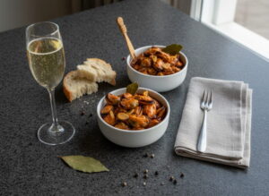 Mejillones en escabeche in weißer Keramikschale, Muscheln in Pimentón-Marinade glänzend, Knoblauch und Lorbeer sichtbar, Sektglas mit perlenden Albariño, galicischer Granit, atlantisches Abendlicht, Tablescape