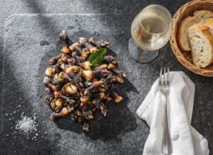 Percebes al natural auf dunklem Holzbrett von oben, Entenmuscheln glänzend mit cremweißem Fleisch, Meersalz daneben, Weißweinglas stark beschlagen, dunkelgrauer Granit, diffuses Atlantiklicht, Overhead